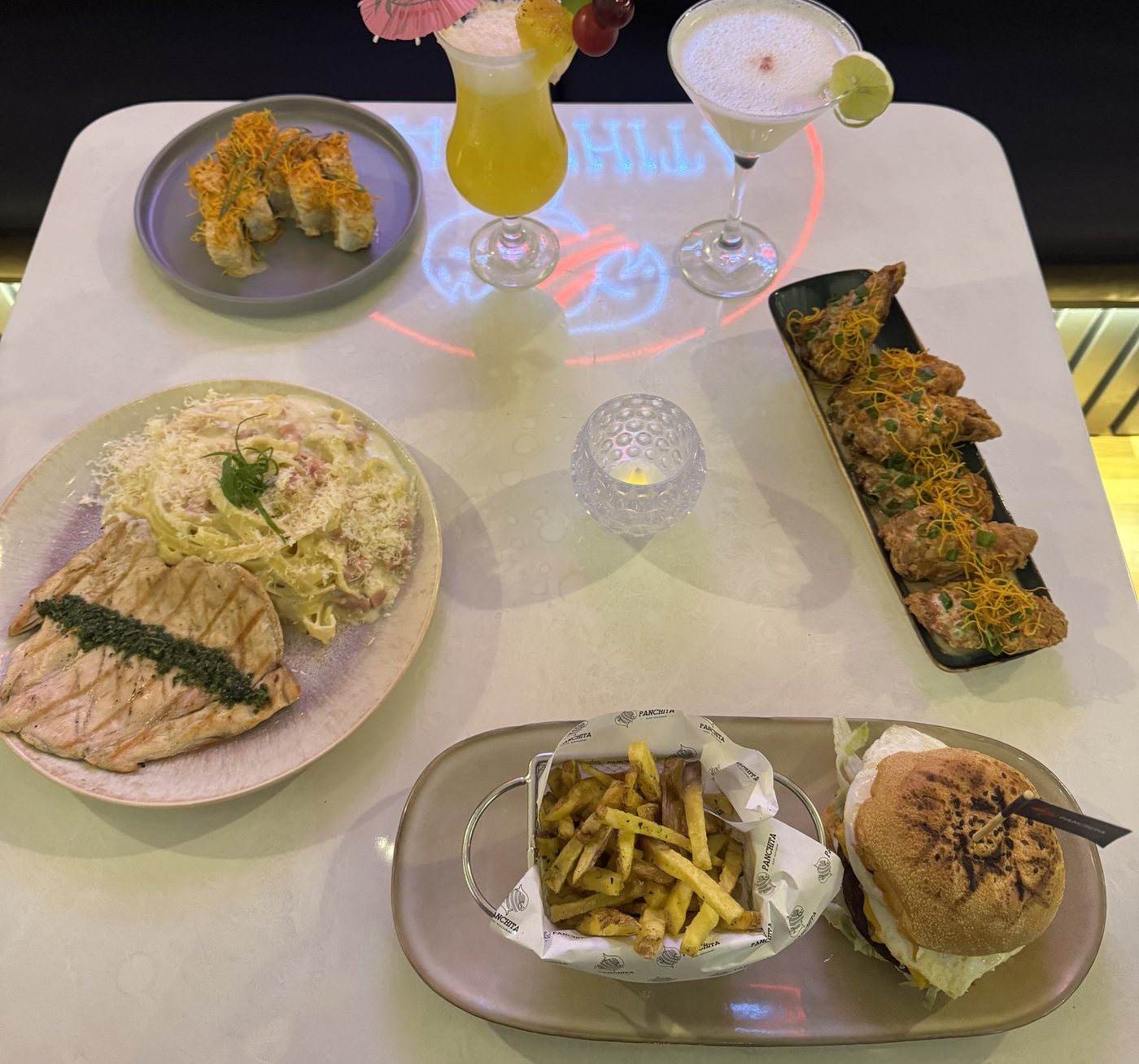 Mesa de Panchita servida con makis, pastas, alitas, hamburguesa y cócteles, con el logo de neón reflejado